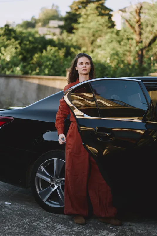 Young woman exits a sleek black car in a long red dress, standing on a sunlit street with green trees in the background, confident and poised, capturing a candid street style moment