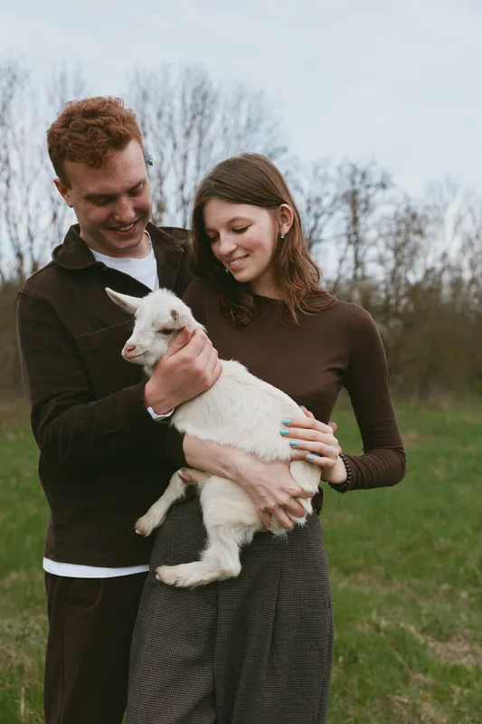 Joyful couple outdoors, holding a goat kid, smiling softly, sunlit moment, casual clothes, calm breeze, natural colors, rural field backdrop, warm connection, candid lifestyle scene, simple