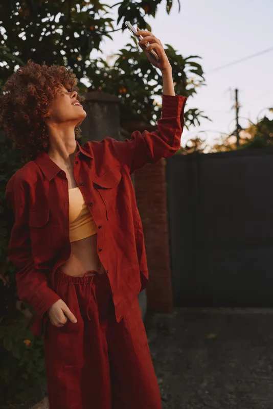 Cheerful woman outdoors, wearing red outfit, reaching toward a branch, sunlit moment, relaxed pose, natural colors, lush greenery background, soft breeze today