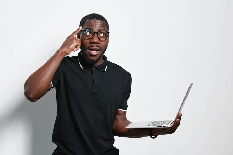 African American man with dark skin and glasses holding laptop and pointing to head thinking about technology and ideas in casual black polo shirt on white background.