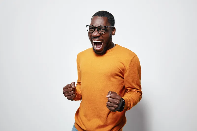 African American man with dark skin wearing glasses and orange sweater expressing excitement with clenched fists and open mouth on white background.
