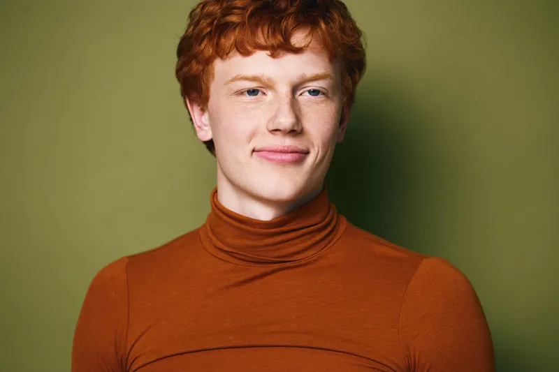 Young man with red hair and freckles wearing orange turtleneck standing against olive green background, portrait with natural expression and clear skin tone in studio lighting.