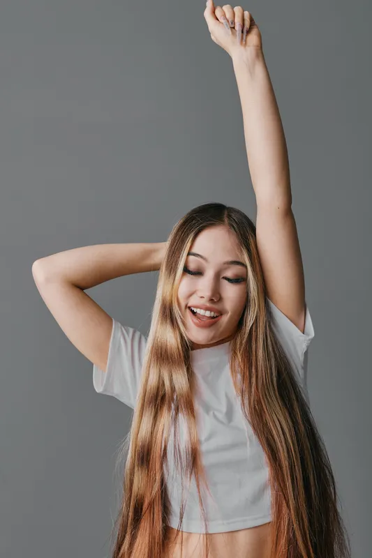 Young woman with long hair smiling and stretching arms wearing white crop top on neutral background studio shot. Happy Asian female with raised hand and closed eyes in casual outfit.