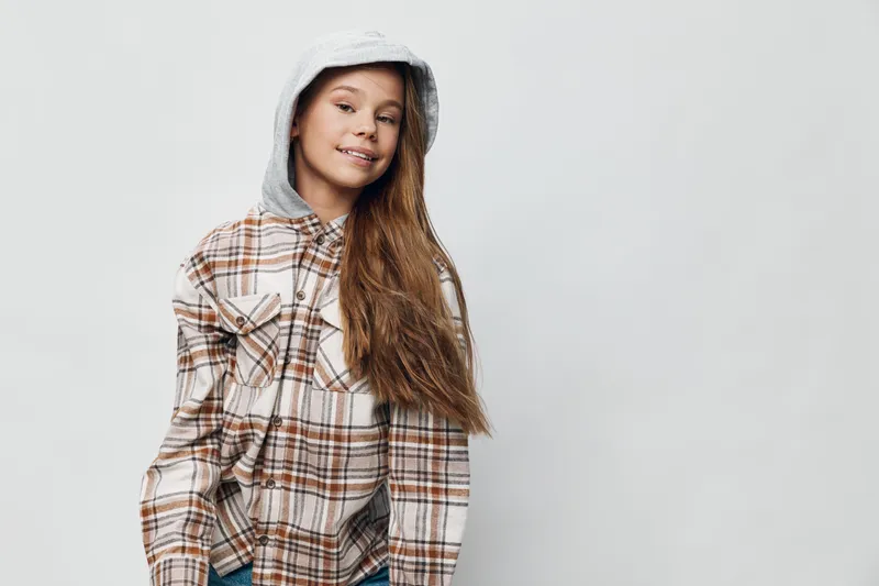 Teenage girl with long hair wearing a hooded sweatshirt and plaid shirt, smiling and posing against a light background for casual style and youth fashion portrait.