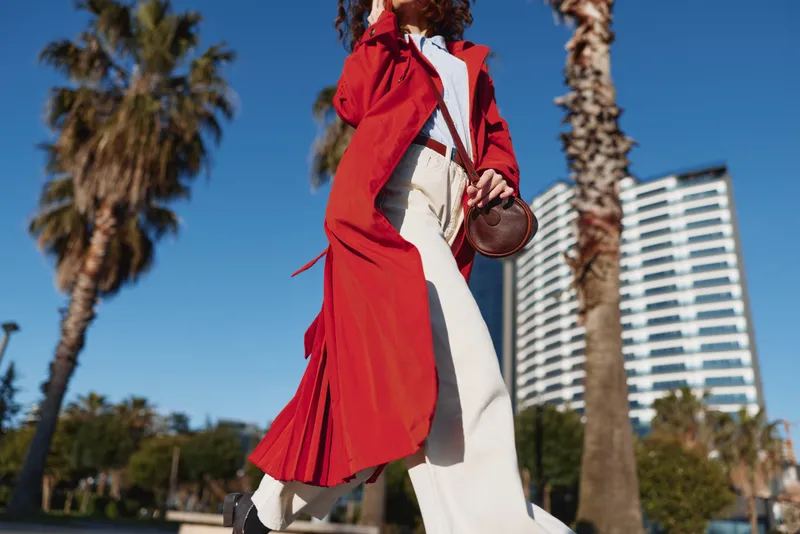 A stylish woman in a bright red trench coat strides along a palm lined street under a clear blue sky, an urban backdrop framing her confident pose.