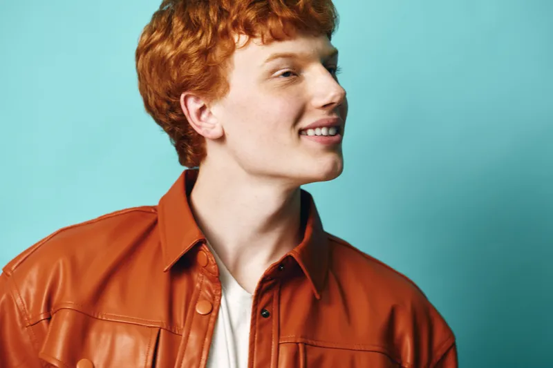 Young man with red hair and freckles smiling wearing brown leather jacket with white shirt on blue background. Portrait of cheerful male model in casual outfit looking sideways.