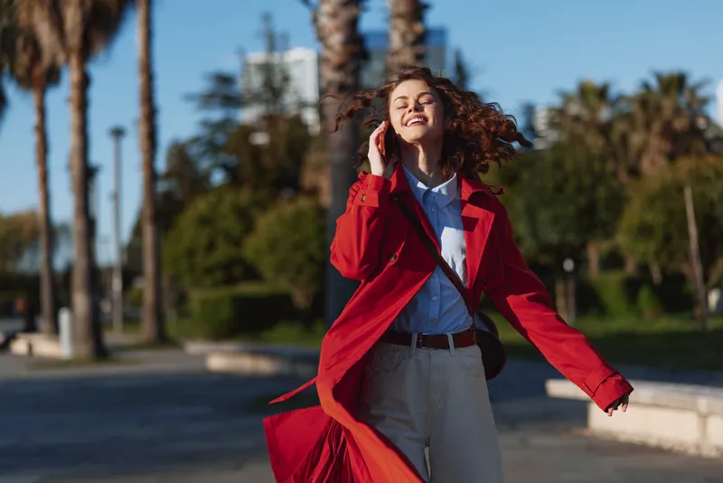 Young woman with curly hair joyfully walks outdoors wearing a red coat and white shirt under bright sunlight on a clear day.