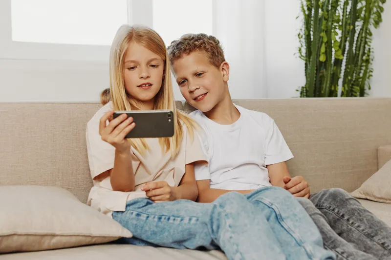 Two children sitting on the sofa taking a selfie with smartphone, smiling and enjoying casual moment in bright living room. Concept of family, childhood and togetherness.