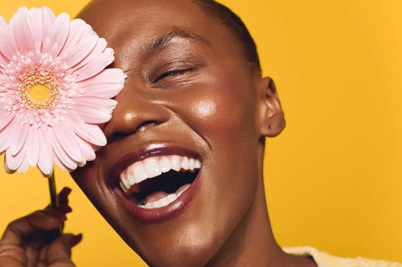 Joyful African woman with dark skin laughing and holding a pink flower near her eye. Bright yellow background highlights her natural beauty and positive mood in a close-up portrait.