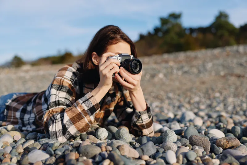 Photographer lying on a pebble beach, focused on capturing shots with a camera, dressed in a plaid jacket, wind in hair, natural light enhances texture and a calm outdoor mood