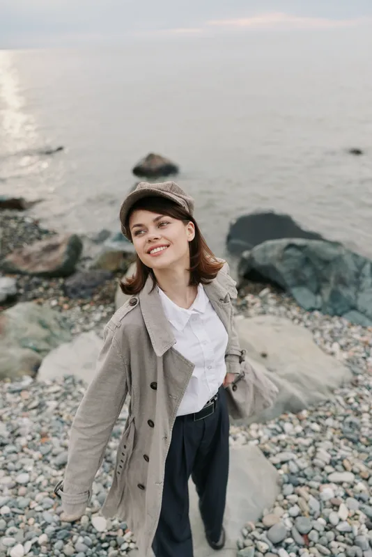Young woman wearing coat and hat stands on rocky beach near sea, smiling and looking away. Casual autumn outfit, outdoor leisure, and natural coastal landscape concept.