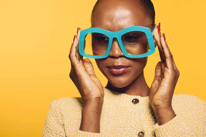 Close-up of a confident dark skinned African woman wearing oversized blue glasses and a beige sweater against a bright yellow background. Concept of fashion, style, and beauty.