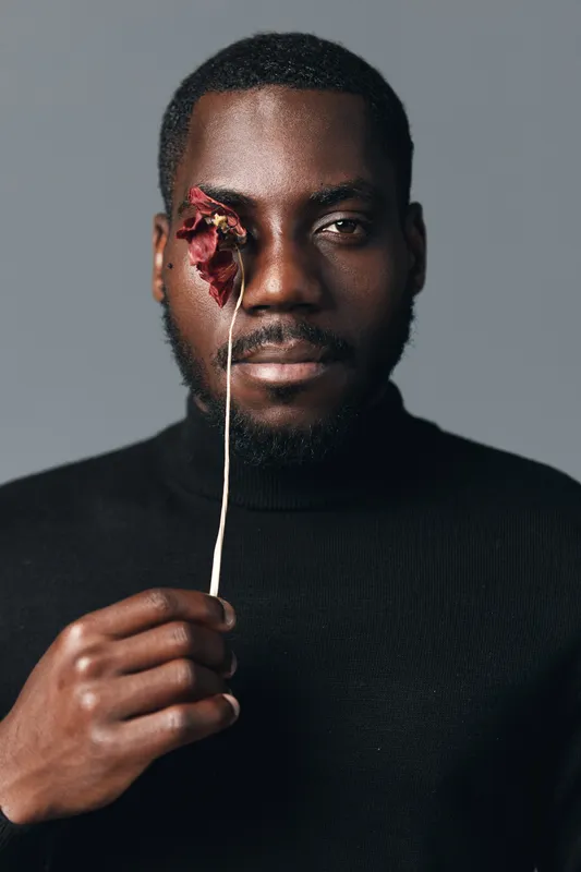 Creative conceptual portrait of African man holding dried flower over eye in visual style with dark skin and black turtleneck on gray background for artistic expression.