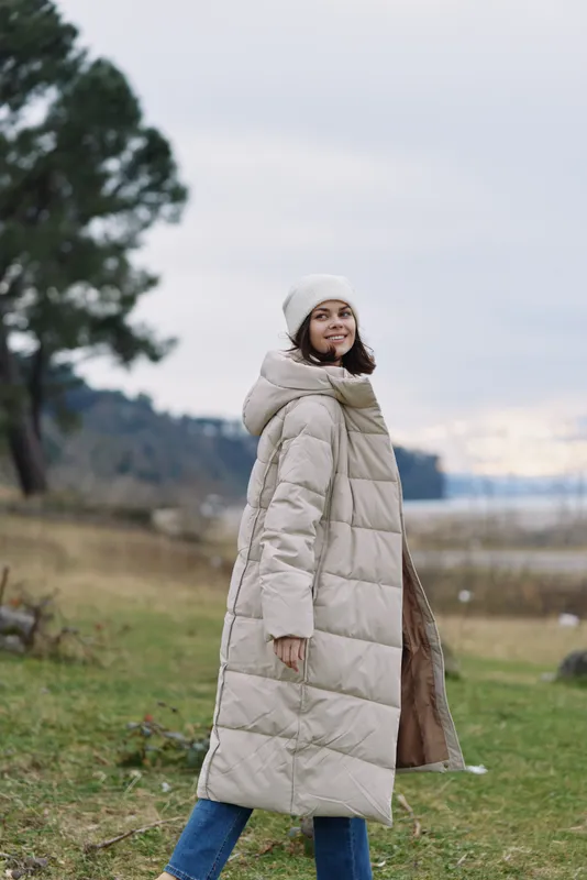 Winter woman in a beige puffer coat walking outdoors by a lake, wearing a beanie, showcasing cozy and stylish cold weather style, vibe.