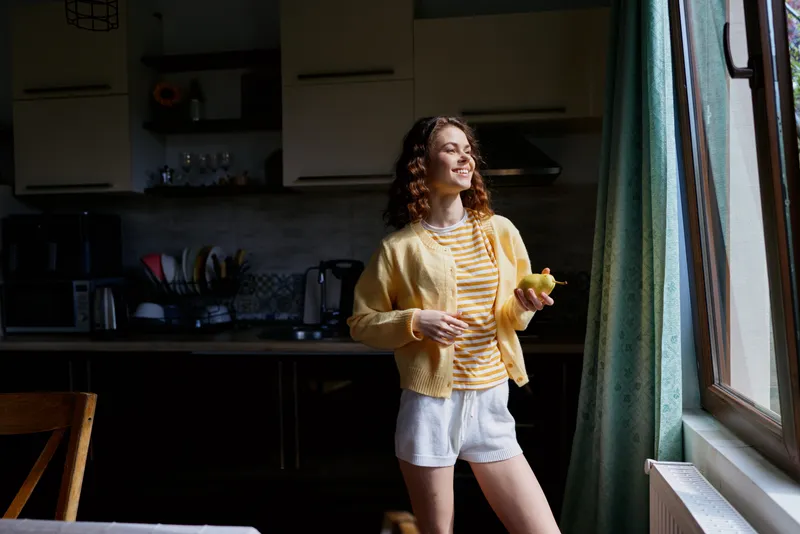 Smiling woman with curly hair standing in a cozy kitchen, wearing striped shirt and yellow cardigan. Casual home lifestyle scene focused on everyday comfort and warm natural light.