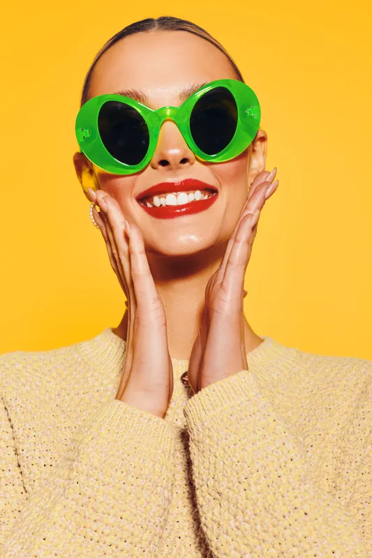 Smiling woman with bright red lipstick wearing large green sunglasses and a yellow sweater poses against a vibrant yellow background. Concept of summer fashion and joyful mood.