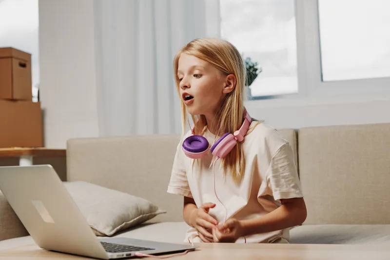 Young girl with headphones sings passionately in front of laptop at home. Natural light from window creates cozy atmosphere. Concept of online singing, music practice, and childhood creativity.