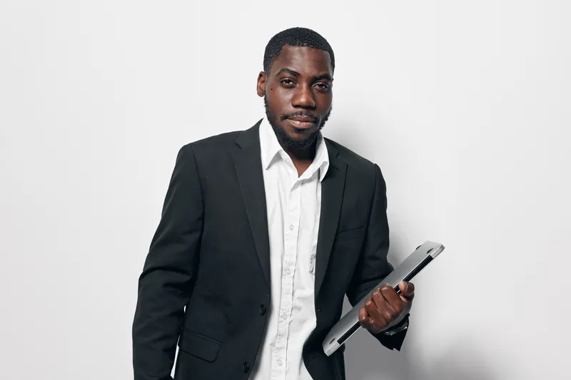 African American man with dark skin wearing black suit and white shirt holding laptop standing against white background looking confident and professional.