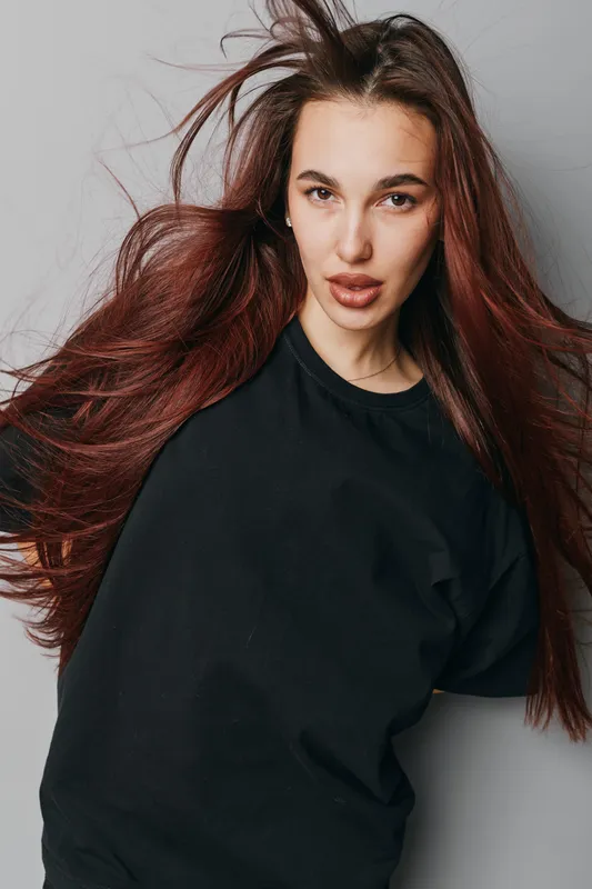 Woman with long flowing hair and natural makeup posing in black shirt against gray background. Female model with confident expression and casual style in studio portrait photography.