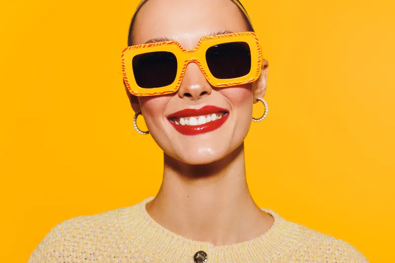 Smiling woman with bright red lipstick wearing large yellow sunglasses and hoop earrings on yellow background. Concept of fashion, summer style, and confident beauty.