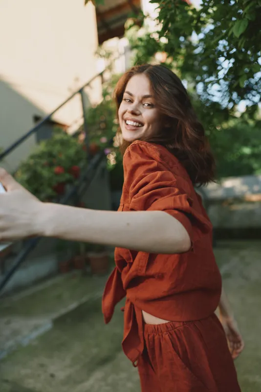 Young woman wearing a rust red outfit twists joyfully outdoors, smiling at the camera, surrounded by greenery and sunlit scenery, creating a bright lifestyle moment perfect for fashion campaigns
