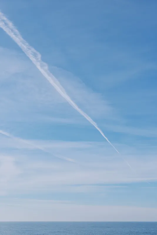 Sky and ocean stretch under a clear blue sky with a diagonal contrail, calm horizon on open water, conveying serenity, vastness and peaceful travel mood.