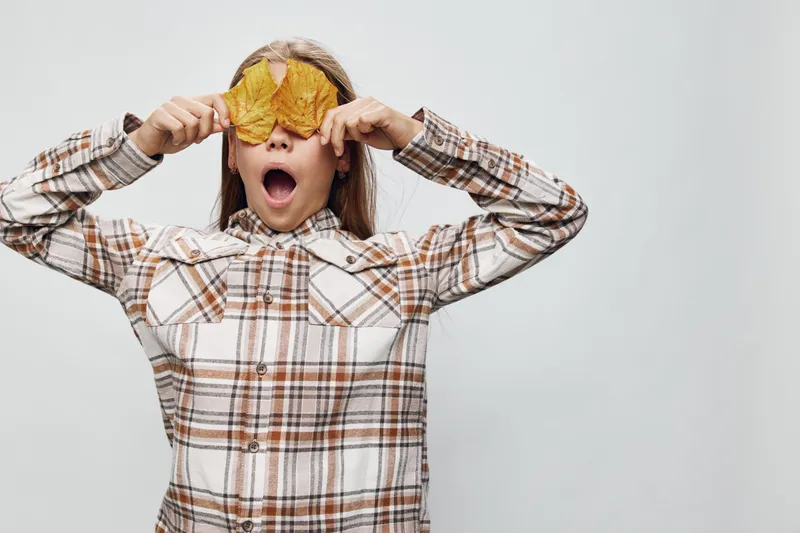 Portrait of surprised girl with long hair wearing plaid shirt holding yellow autumn leaves over eyes on white background for seasonal concept and playful mood.