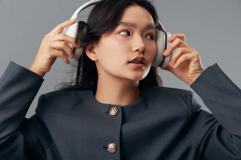 Woman with long black hair wearing grey jacket adjusting white headphones while looking to side in studio. Portrait of young female with technology accessory and casual style.