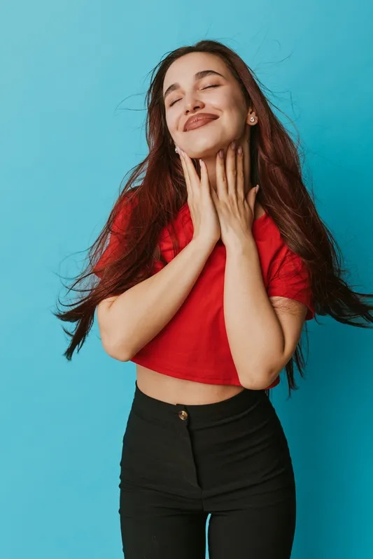Young woman with long hair wearing red cropped top and black pants posing with closed eyes and smiling on blue background. Beauty, happiness, casual style and confident female portrait.
