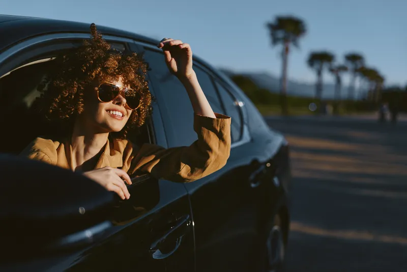 Cheerful woman in a sleek car, sunlit outdoors, smile with sunglasses, casual lifestyle, breeze in hair, vintage color tones, vibrant, candid moment of lifestyle and freedom on the road