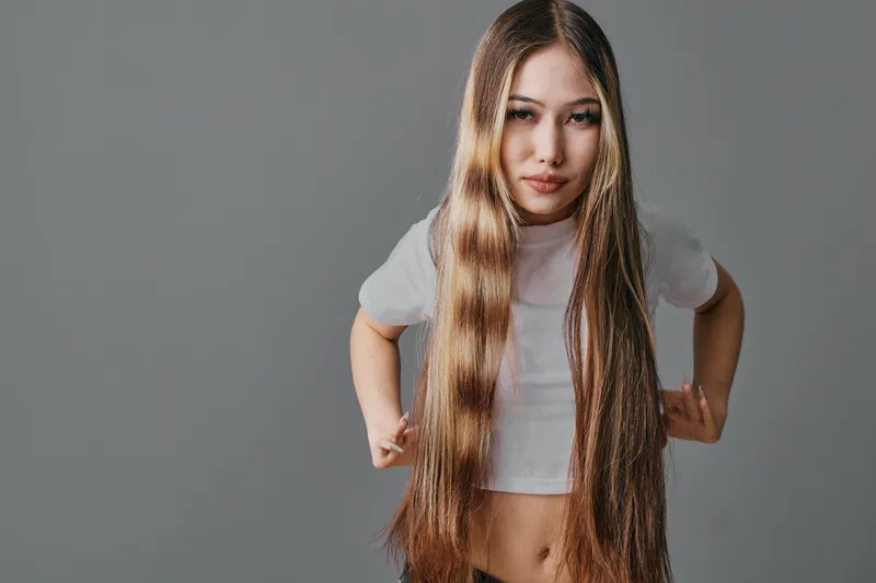 Young woman with extremely long straight hair wearing white crop top posing confidently on grey background with subtle expression and hands on hips in studio light.