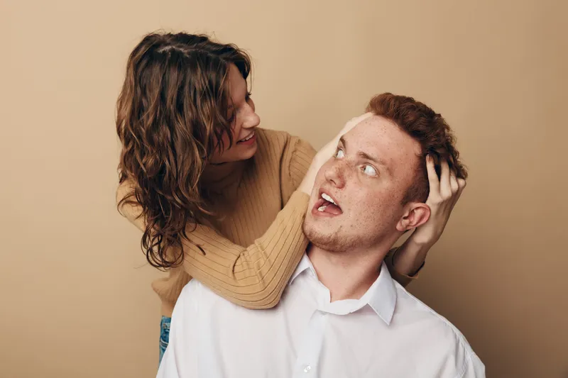 Young woman with curly hair playfully holding surprised redhead man by head. Casual interaction between friends or couple on beige background. Concept of fun, friendship, and emotions.