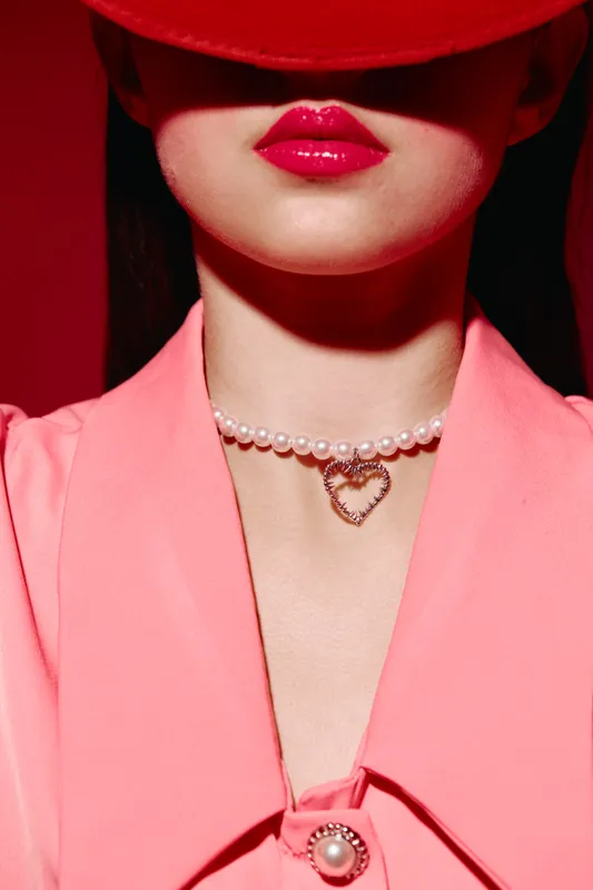 Close-up of a fashionable woman wearing a bright pink blazer and hat, with glossy red lips and a pearl necklace featuring a heart-shaped pendant. Creative beauty fashion concept.