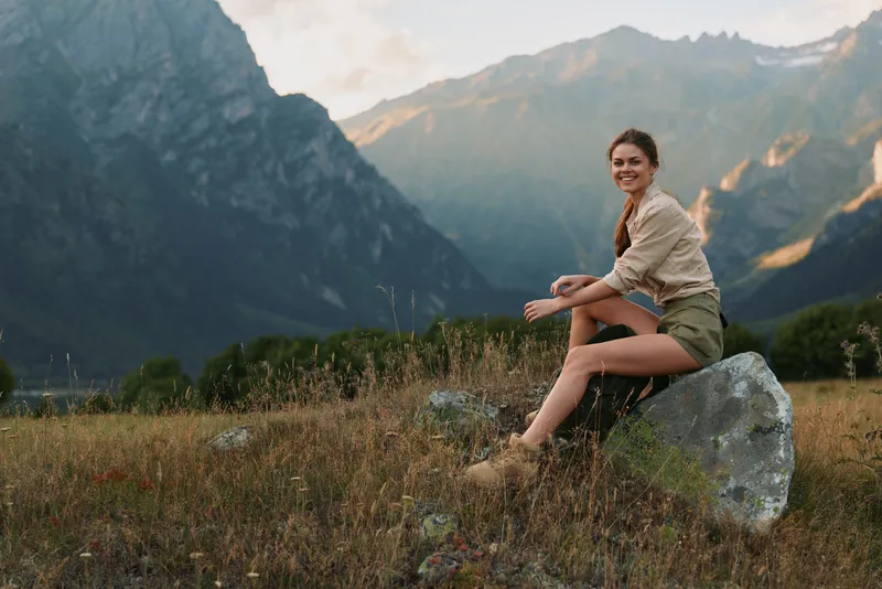 Woman sitting on a rock in a mountain meadow, outdoor lifestyle shot, nature scene with mountains in the background, tranquil yet adventurous mood and lighting highlighting grass and rock too