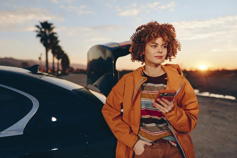 Woman lifestyle on roadside at sunset, standing by parked car, curly hair, wearing orange jacket, holding smartphone and smiling at camera, casual travel mood, warm golden light, palm trees