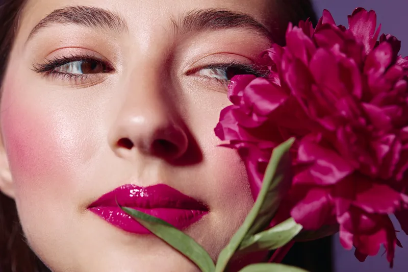 Close-up portrait of a woman with glossy magenta lipstick and shimmering eyeshadow holding a deep pink flower near her cheek. Studio shot with soft lighting reveals beauty and fashion themes.
