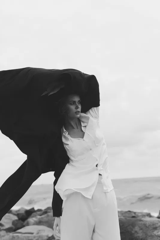 Fashion silhouette on a windy beach features a flowing coat over a white shirt, capturing movement and mood in a monochrome coastal scene by the sea, with a stark sky and rugged shoreline for