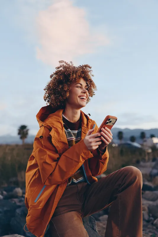 Woman with curly hair, smiling outdoors, wearing a jacket, holding a smartphone while sitting in a field, warm sky and distant mountains, relaxed lifestyle, casual travel moment