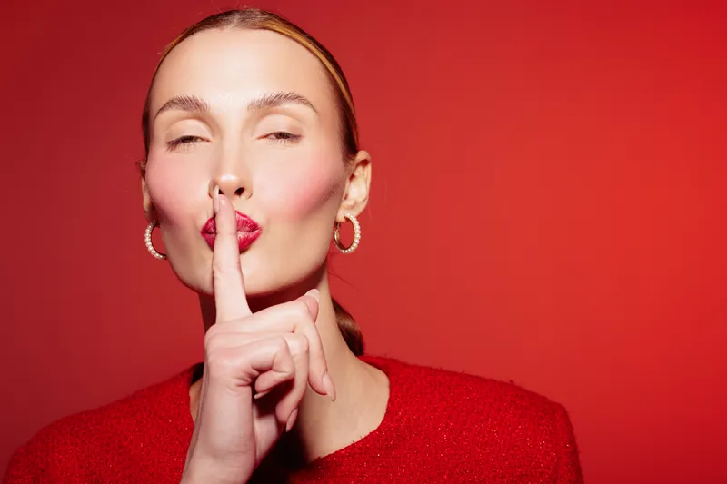Young woman with red lipstick and earrings makes silence gesture with finger on lips. Close-up portrait on red background. Concept of secrecy, quiet, and calm expression.