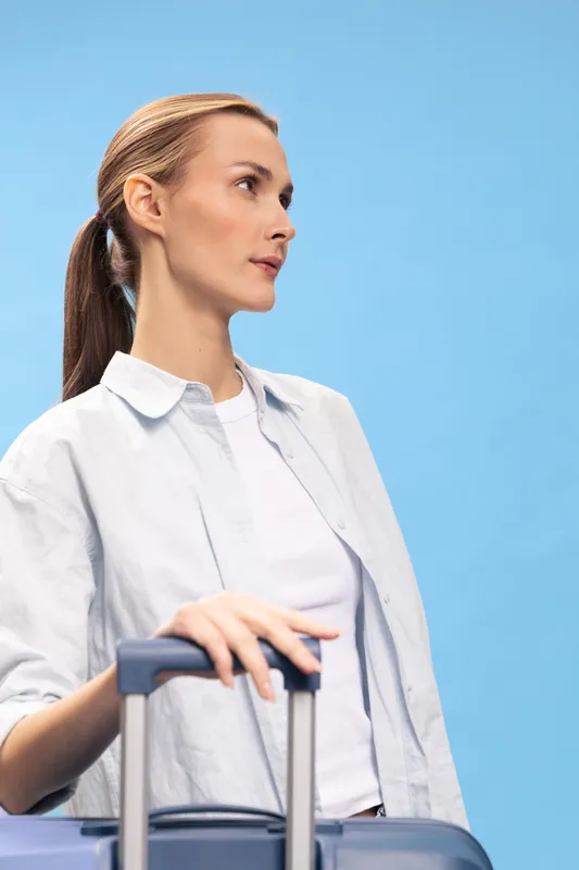 Young woman with a ponytail stands holding a suitcase handle against a blue background. She looks thoughtfully to the side, wearing a white shirt and light blouse. Travel concept.