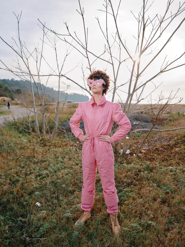 Fashionable woman in a pink jumpsuit stands confidently in an open field with bare trees, warm sun, blue sky, and a rustic landscape, conveying bold style and outdoor charm.