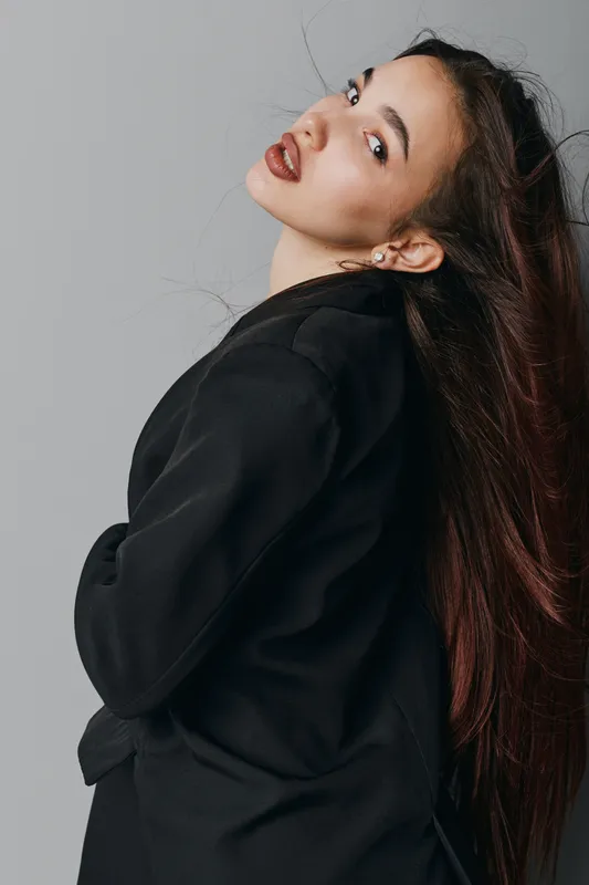 Woman with long hair and black jacket posing sideways against gray background. Female model with minimal makeup and earrings looking over shoulder with confident expression.