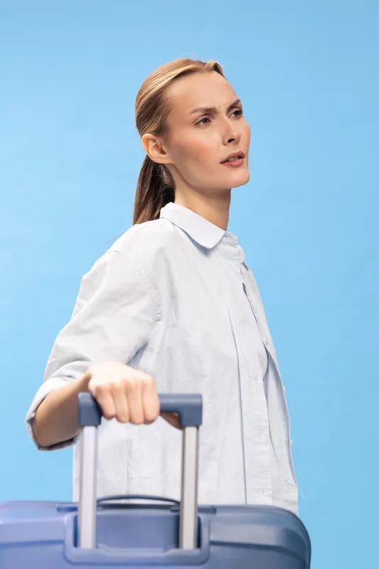 Young woman in white shirt holds suitcase handle, looking away on blue background. Concept for travel, planning, journey, and vacation preparation.