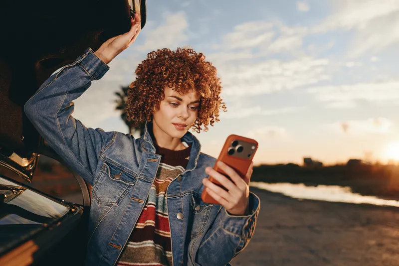 Woman with curly hair, in a denim jacket, holds a smartphone for a selfie, outdoors at sunset, golden light, casual lifestyle portrait, relaxed smile, beach road background