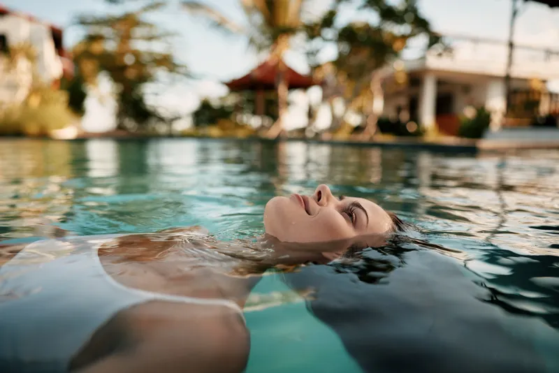 A person floats peacefully in a clear pool, head tilted back, eyes closed, enjoying a tranquil moment by a resort as sunlight glints on the water, conveying vacation lifestyle.