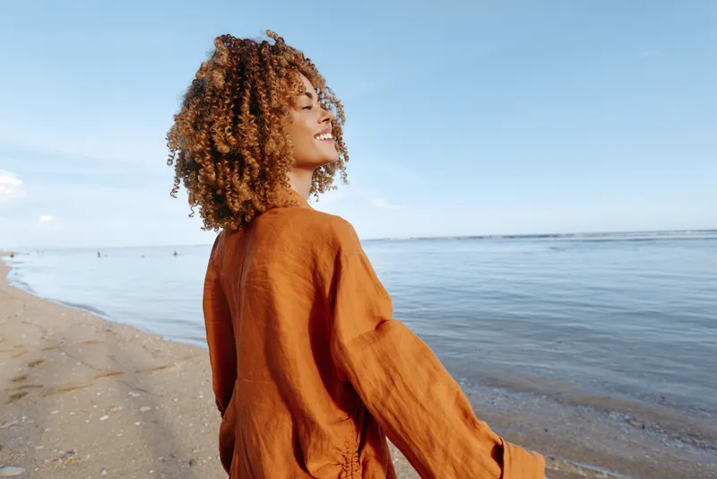 Woman with curly hair in profile on a sandy beach, wearing a warm orange sweater. Relaxation and nature mood by the ocean under soft sunlight, summer outdoors.