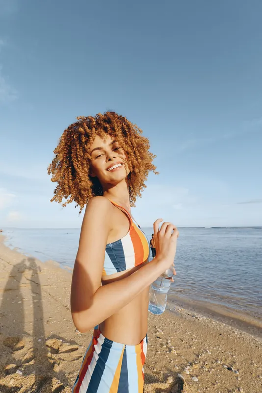 Woman with curly hair standing on beach by ocean, smiling in warm sunlight, wearing striped swimsuit, relaxation posture, summer seaside mood, gentle breeze, clear sky background, calm