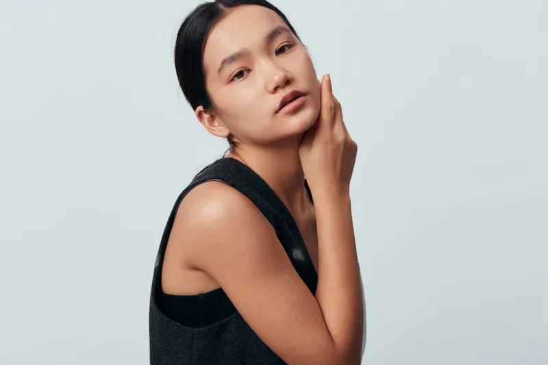 Young woman with smooth clear skin and natural makeup posing in sleeveless top against light background. Female model with hand on face expressing calm and elegance in studio.