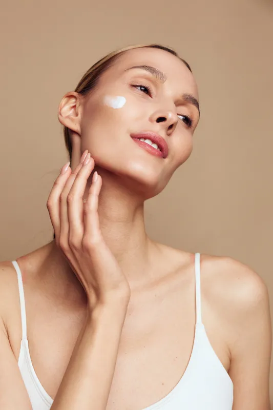 Young woman applies moisturizing cream on her face gently touching skin with hand, showing skincare routine and natural beauty on beige background in white top.