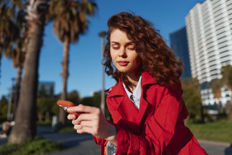 Young woman with a red jacket focuses on her phone in an urban park, with palm trees and tall buildings in the background, a candid moment of everyday connection.
